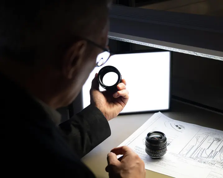 A man inspects a camera lens, technical drawings lie on the table.