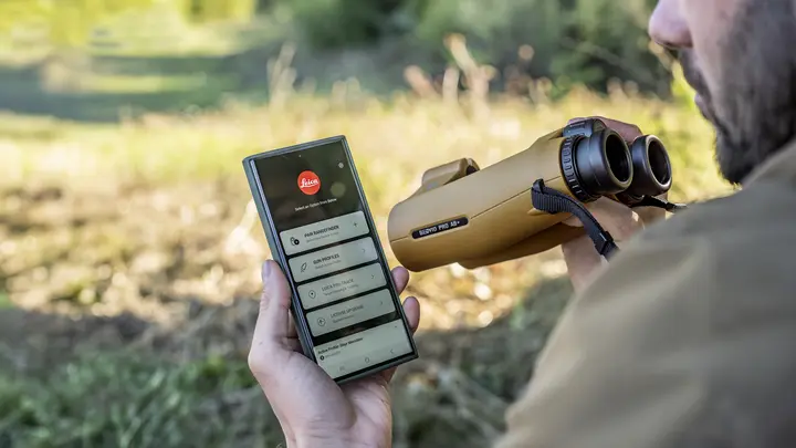 A mobile phone held by a hunter, opened with Leica Ballistics App and Leica Geovid Pro AB+ in the foreground, a meadow can be seen in the background.