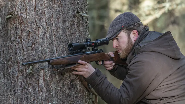 The Leica Fortis 6 1.8-12x42i is in focus while a hunter leans against a tree with his weapon and looks through the scope