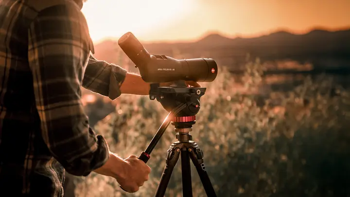 The Leica APO Televid is in the foreground while being operated by a man in a lumberjack shirt. In the background is a picturesque landscape at sunset