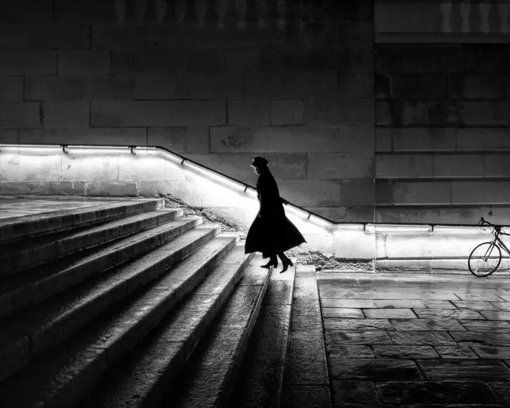 A woman in a long coat is walking up a staircase at night. The banister is illuminated.