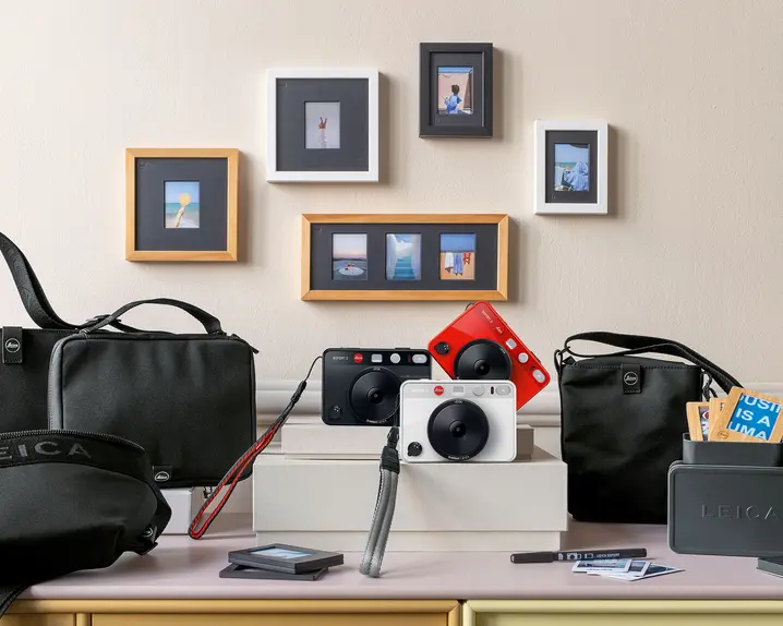 Leica cameras and accessories on a table, with framed photos on the wall in the background.