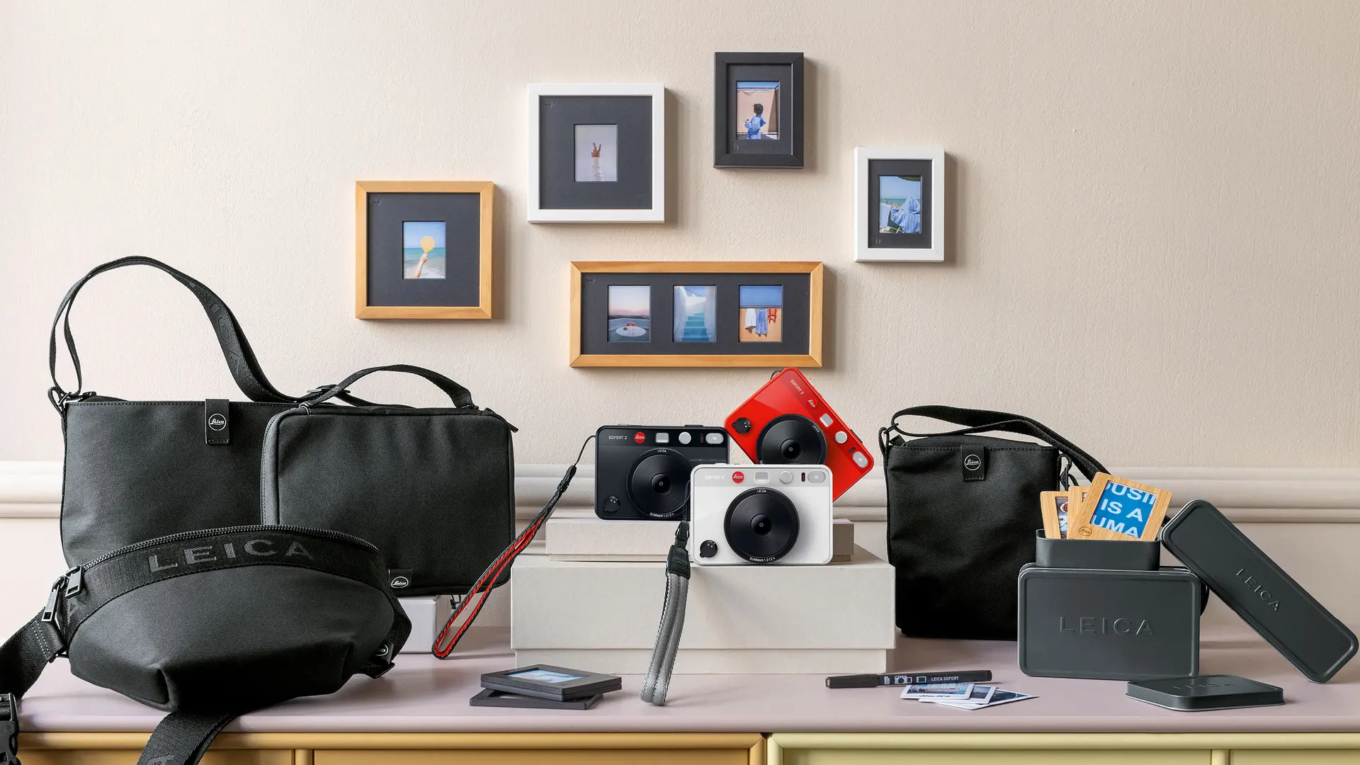 Leica cameras and accessories on a table, with framed photos on the wall in the background.