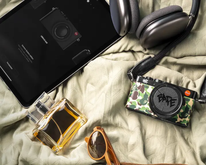 A camera, tablet, headphones, sunglasses, perfume and wallet laying on a bedcover.