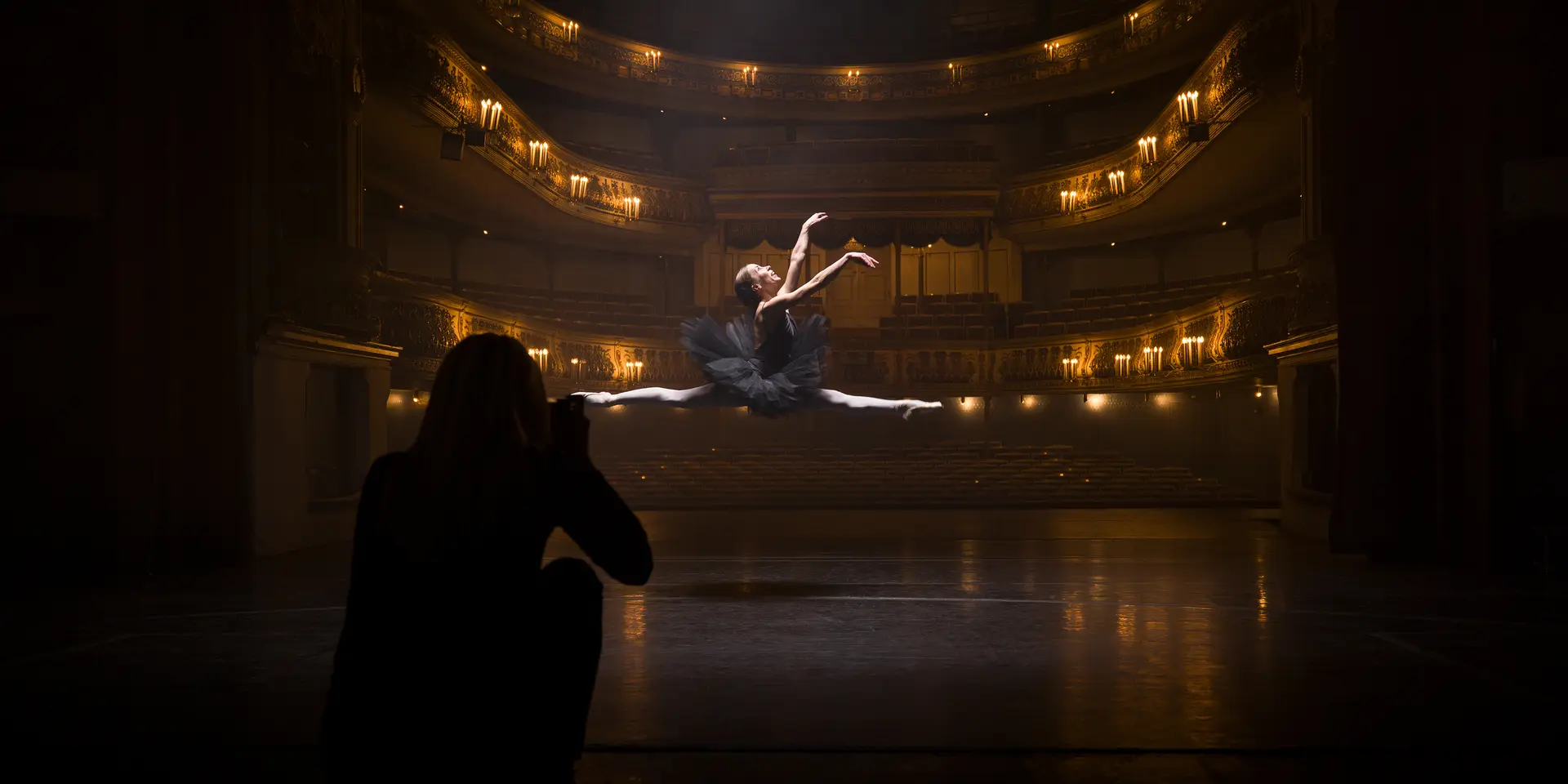 A person in the shade takes a picture of a ballerina jumping in an illuminated theatre.
