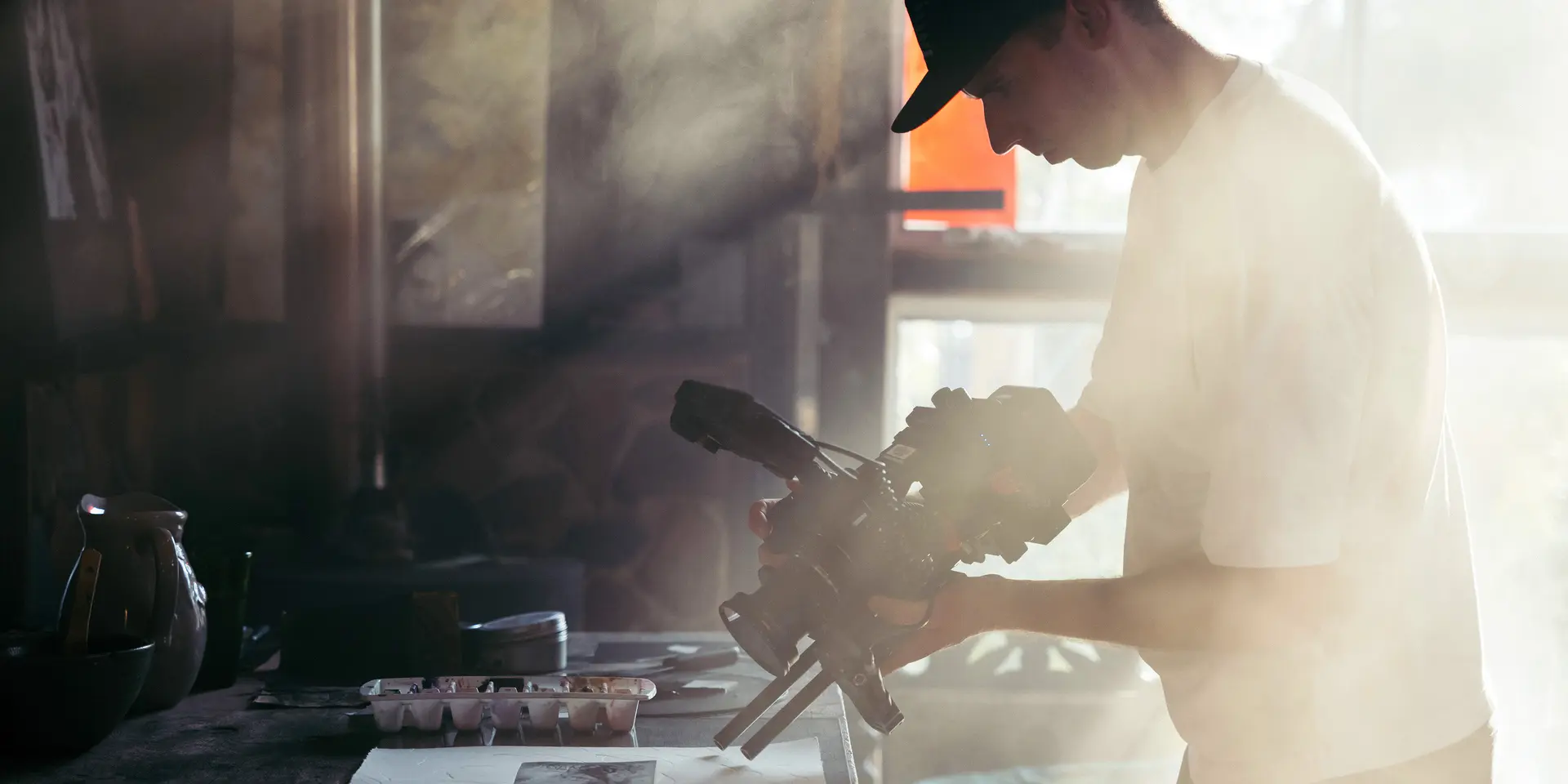 Person holds camera over a table with art materials, warm sunlight falls through window.