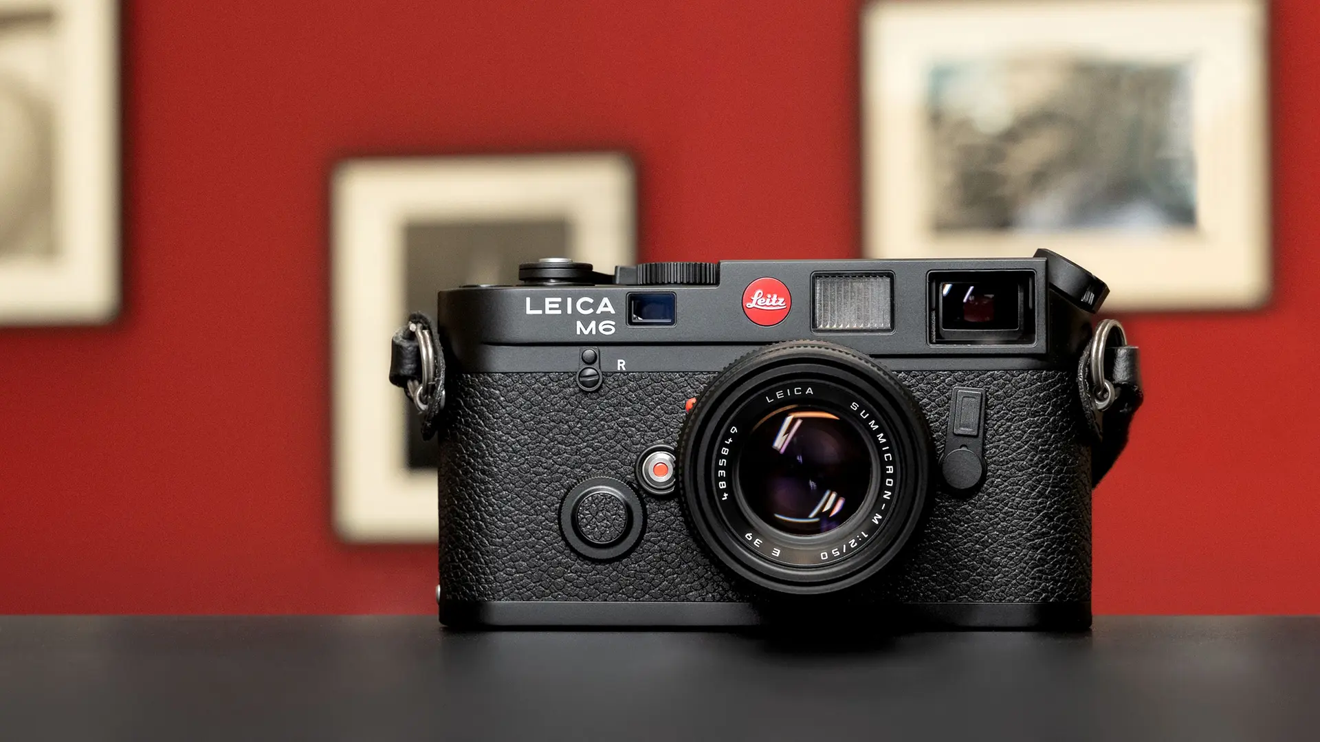 Leica camera stands on a table, framed photos hang on the wall in the background.