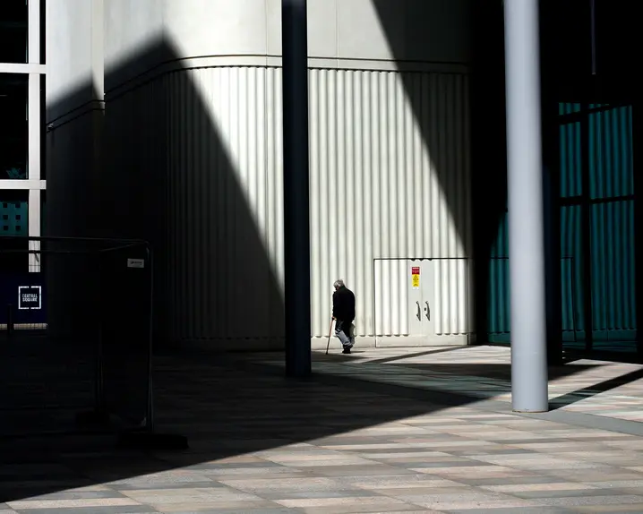An old man walks in front of a building with shadows beside him.