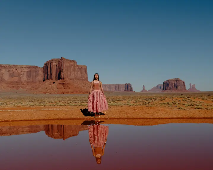 Woman standing in a desert landscape with red rock formations in the background, the image is reflected in the water in front of her