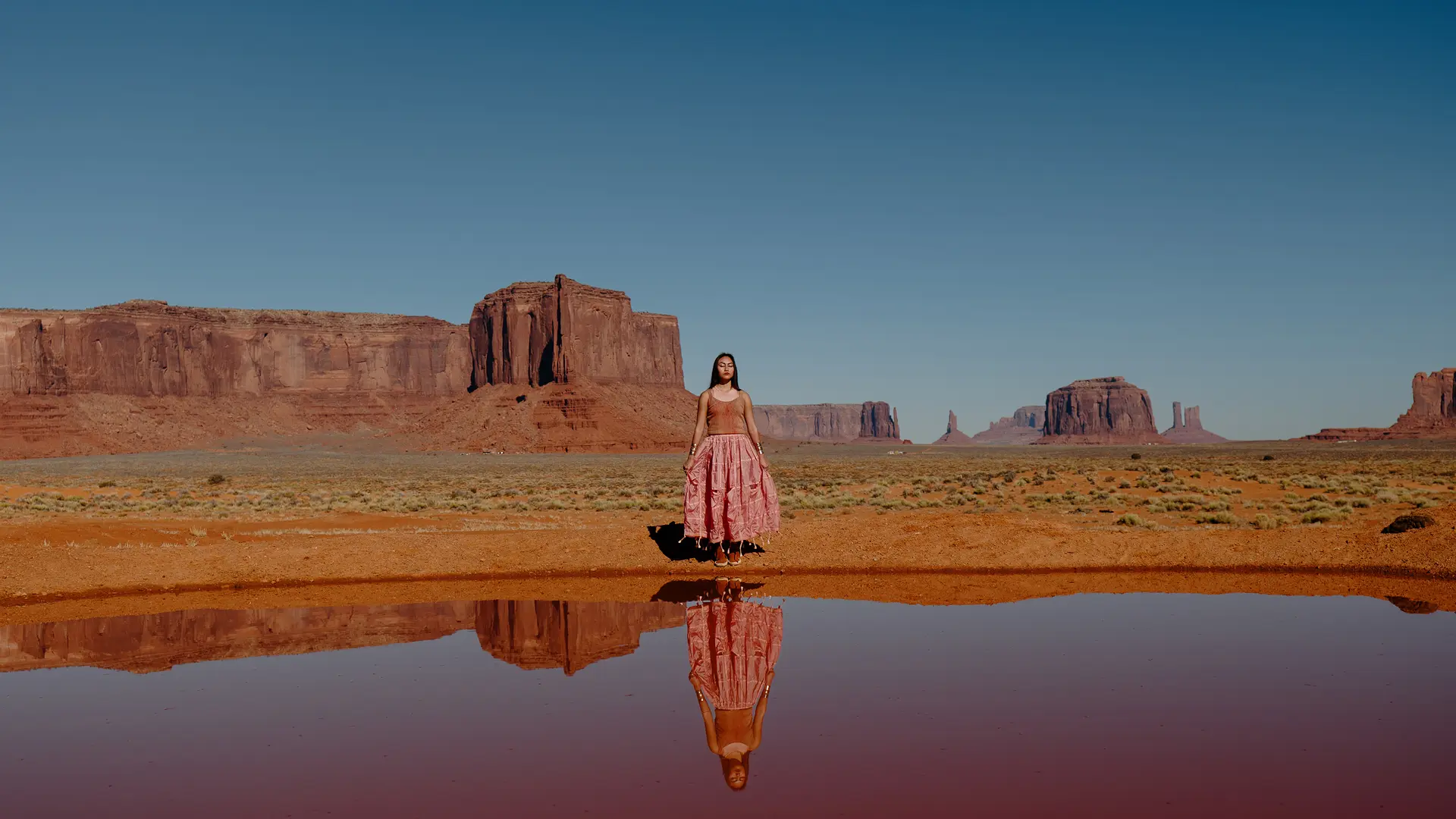 Woman standing in a desert landscape with red rock formations in the background, the image is reflected in the water in front of her