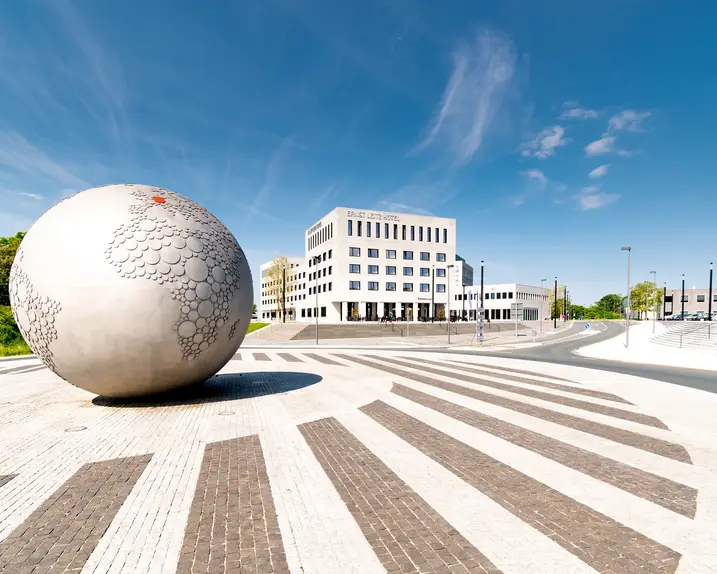Large stone globe on a square in front of a hotel and parking lot under a blue sky.